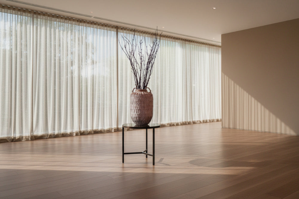A large textured pink ceramic vase with two handles, filled with dried branches, displayed on a small round table in a modern, sunlit room with sheer curtains.