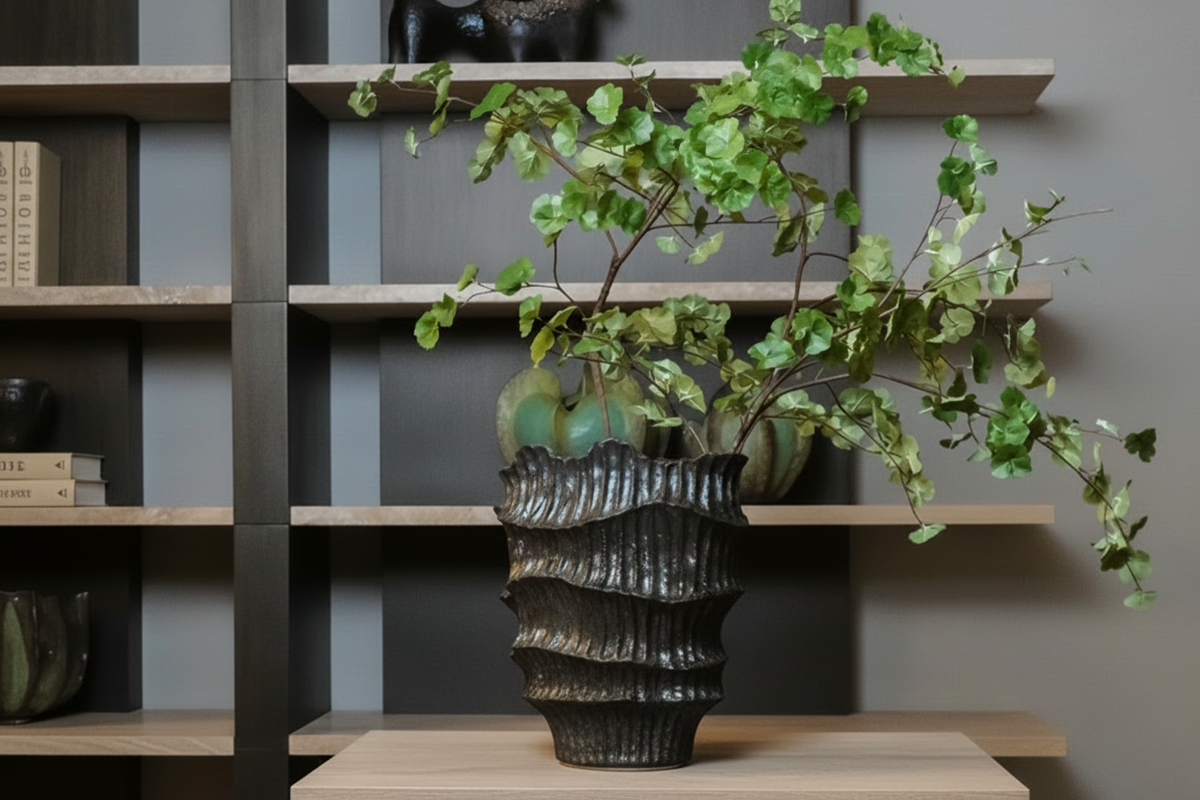 A green trailing houseplant in a textured dark gray ceramic pot, displayed on a wooden shelf against a gray wall.